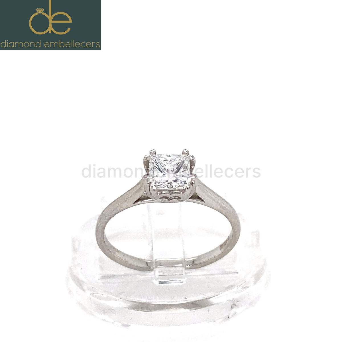 A princess-cut diamond solitaire ring in a platinum band, displayed on a clear acrylic stand against a white background. 