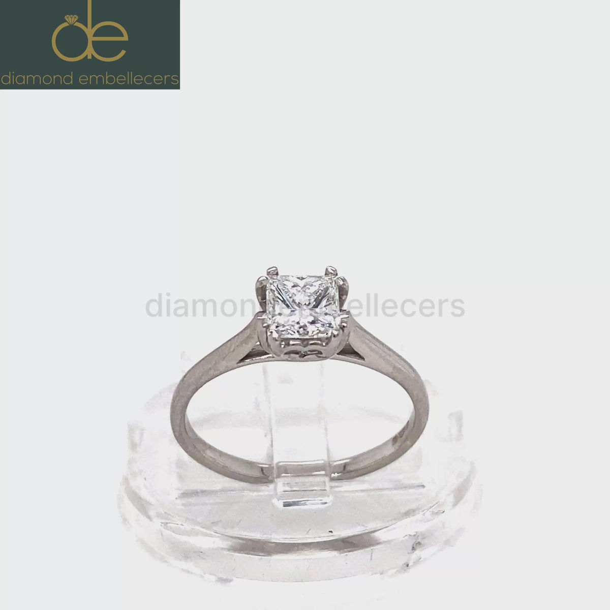 A princess-cut diamond solitaire ring in a platinum band, displayed on a clear acrylic stand against a white background. 